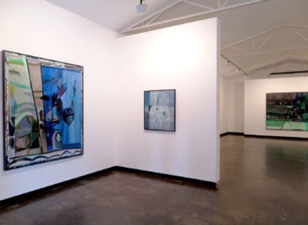 Interior view of a contemporary art gallery with white partition walls, polished dark concrete flooring, exposed white ceiling structure and track lighting. Several framed abstract artworks are displayed throughout the open exhibition space.