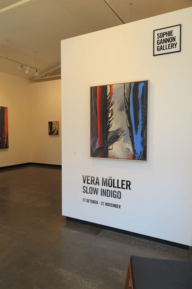 Interior view of a contemporary art gallery with white partition walls displaying framed abstract paintings. A prominent exhibition panel identifies the Sophie Gannon Gallery and promotes Vera Möller’s “Slow Indigo” exhibition. The space has polished concrete flooring, track lighting, exposed ceiling services, and a minimal industrial gallery atmosphere.