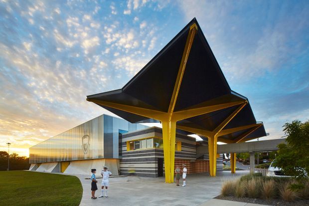 Exterior architectural view of a contemporary institutional or commercial building at sunset. The structure features dramatic angular black tensile or folded canopy roofs supported by vivid yellow structural columns, a glazed and metal-clad facade, striped horizontal wall finishes, and a broad paved entrance plaza with landscaped planting. Several people provide scale.