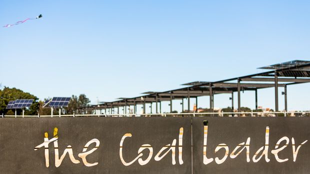 Exterior view of a large steel-framed building or structure under construction, with extensive roof framing, solar panels at left, and a dark wall in the foreground bearing the words “The Coal Loader.” Clear blue sky and a kite are visible above.