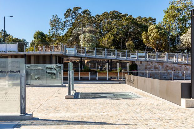 A contemporary low-rise public or commercial building viewed from a broad paved plaza. The structure features extensive glass storefront glazing, dark vertical cladding, exposed concrete elements, pale stone retaining walls, and a raised terrace with metal-and-glass balustrades. Mature trees surround the site, and a glazed information or display pavilion occupies the foreground.