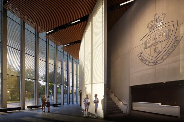 A grand, double-height institutional foyer or reception lobby with full-height glazed curtain walls, vertical blinds, a warm timber slatted ceiling, pale stone or concrete feature walls, and a reception desk beneath a large engraved crest. A staircase rises along the right wall, while several people provide scale. The space has a refined, contemporary civic or educational character and opens visually to landscaped grounds outside.
