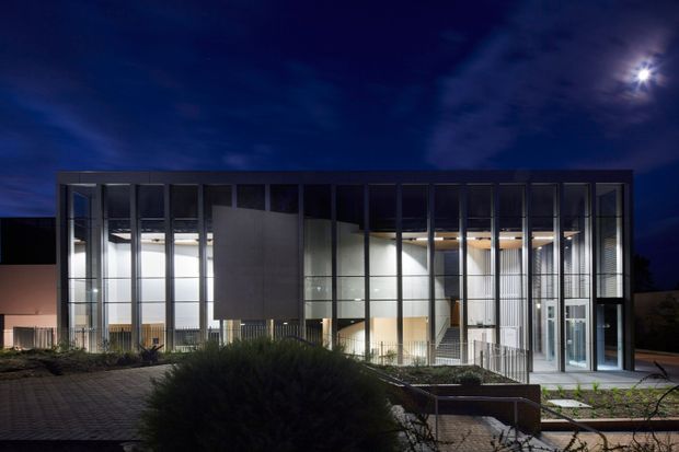Night-time exterior view of a contemporary multi-storey glazed building. The facade is composed of extensive floor-to-ceiling glass, dark vertical metal framing, solid dark panels, and warm illuminated interior spaces. A paved forecourt with planted landscaping and steps leads toward the entrance, while exposed interior stairs and timber-toned ceiling elements are visible through the glazing.