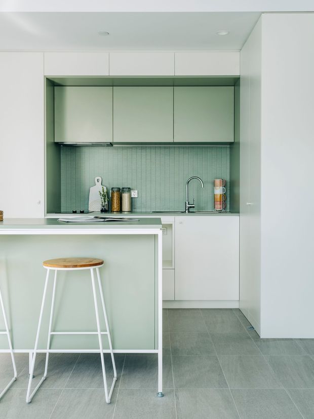 A compact, minimalist kitchen with pale mint-green and white cabinetry, a matching small-format tiled splashback, and a peninsula island with a light wood-topped white stool. The kitchen has integrated upper and lower joinery, a stainless or chrome gooseneck tap, recessed ceiling lights, and large grey floor tiles. The overall composition is clean, light, and highly streamlined.