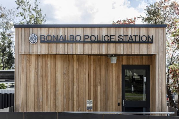 Exterior view of a compact contemporary police station clad in vertical timber boards. The facade features prominent dark lettering reading “BONALBO POLICE STATION,” a police emblem, a recessed entry canopy, and a dark-framed glazed door, with a simple flat roofline and surrounding trees.