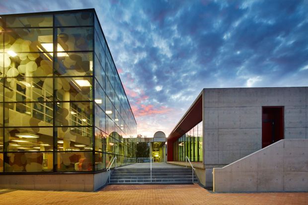Exterior architectural view of a contemporary institutional or commercial building at dusk. The composition shows a large transparent glazed volume with dark metal framing and illuminated interiors on the left, connected to a minimalist concrete structure on the right by a recessed entry courtyard and broad steps. The concrete building has a flat roof, a deep projecting canopy, and a dark red doorway. Brick paving, handrails, and dramatic evening clouds complete the scene.