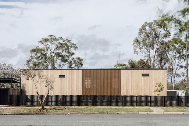 Street-facing view of a low, contemporary single-storey building with a simple rectangular form, pale vertical timber cladding and a darker central timber-clad section. A black vertical-slat fence runs across the frontage, with mature trees and a paved street in the foreground.