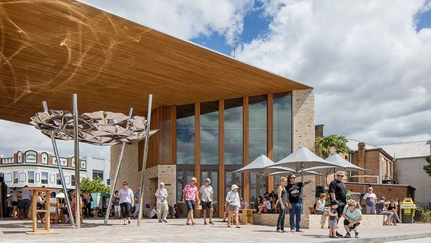 Exterior view of a contemporary public or commercial building with a broad cantilevered timber-clad roof and soffit, tall floor-to-ceiling glazed walls framed in timber, and pale masonry side walls. The building opens onto a paved civic plaza with umbrellas, seating, people, and a sculptural metal canopy or installation. Historic-looking neighbouring buildings are visible around the plaza under a partly cloudy sky.