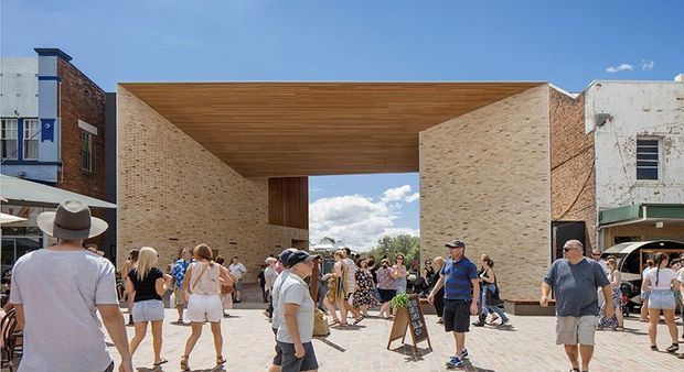 Contemporary urban public building or pavilion framed by two pale brick walls and a broad timber-lined overhead canopy, creating a monumental covered passage or courtyard entrance. Pedestrians fill the paved forecourt, with older brick commercial buildings adjoining on both sides. Bright daytime setting with strong geometric forms and exposed natural timber soffit.