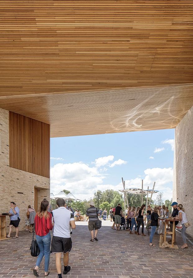 A covered outdoor passage or entry plaza framed by substantial brick walls and a broad timber-lined soffit. The open-sided space leads to a lively landscaped public area, with people gathered around temporary tables and seating. The architecture combines warm horizontal timber cladding overhead and on one wall with textured masonry, creating a sheltered yet airy threshold.