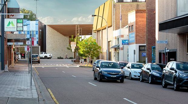 Urban streetscape viewed from the sidewalk, showing a commercial town-centre block with pedestrian paving, parked cars, low- to mid-rise brick and rendered buildings, shopfronts, signage, and a contemporary yellow curved facade element. The scene is a daytime exterior view of a built-up commercial area.