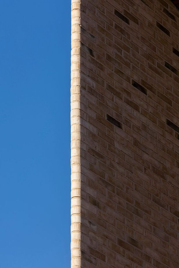 Close-up exterior architectural detail of a tall curved or rounded-corner brick wall against a clear blue sky. The facade uses warm brown masonry with narrow horizontal slit openings, while the corner is highlighted by pale rounded brickwork. No interior spaces are visible.