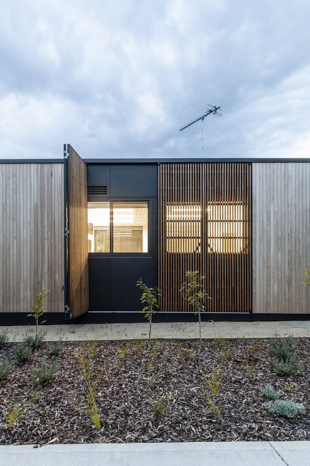 Front-facing exterior view of a low-rise contemporary building with pale vertical timber cladding, dark metal framing, and a prominent vertical timber privacy screen over the glazing. A hinged timber gate or panel is open beside the entrance, revealing a brightly lit interior. The facade sits behind a small landscaped garden bed with young plants and a concrete path; the overcast sky and rooftop antenna are visible above.