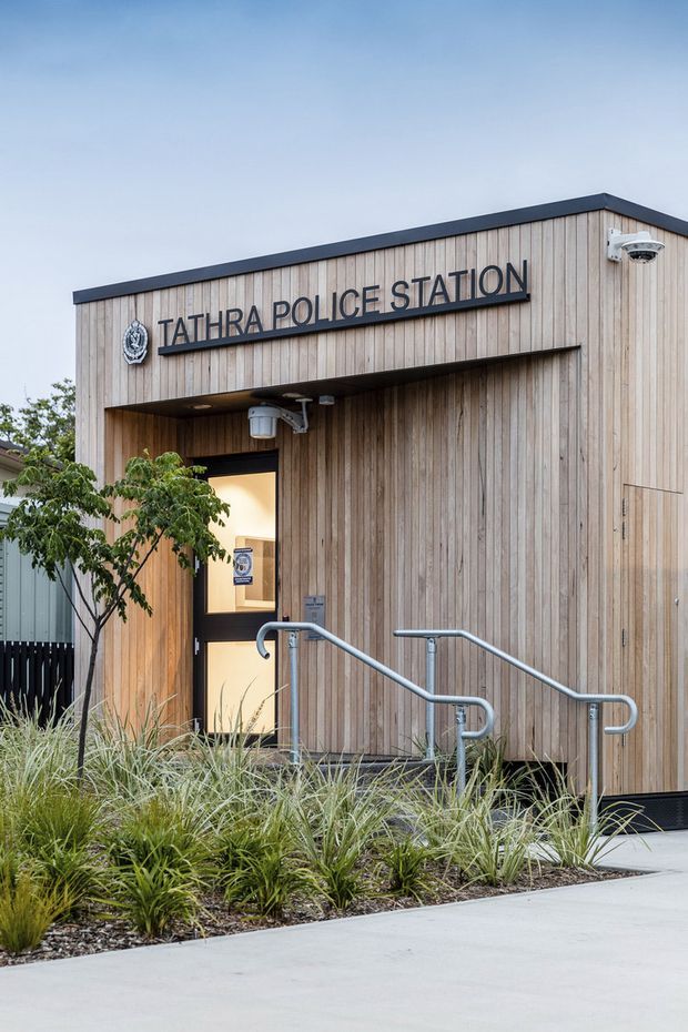 Exterior view of the Tathra Police Station, a small civic building clad in vertical light timber boards. The simple low-pitched roof has dark metal edging, with a recessed illuminated entry, accessibility handrails, security camera, signage, and landscaped ornamental grasses along a concrete approach.
