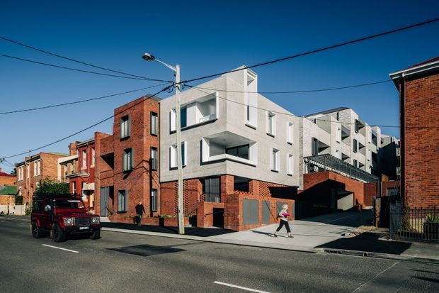 Street-level view of a contemporary multi-storey residential development integrated into an established urban neighbourhood. The building combines crisp white rendered upper levels with exposed red-brick lower walls and adjacent older brick terraces, featuring projecting balconies, recessed glazing, a covered vehicle entry, and a central urban driveway. Overhead utility wires and street infrastructure are visible.