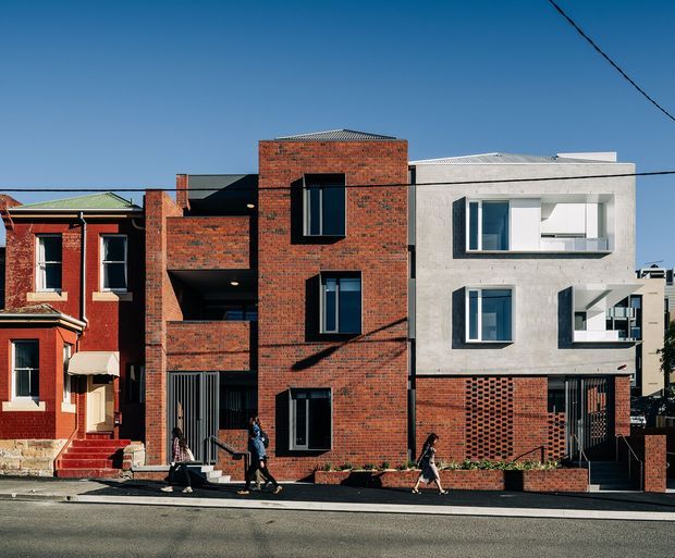 Street-facing view of a row of attached urban dwellings, showing a traditional red-brick residence beside a contemporary multi-storey brick-and-concrete townhouse. The newer buildings feature strong rectangular volumes, recessed balconies, dark-framed windows, patterned brickwork, and a flat or very low-pitched roofline. Pedestrians and street infrastructure are visible in the foreground.