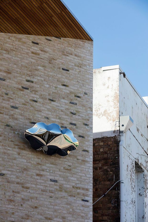 Exterior architectural detail showing a pale exposed-brick wall beneath a steep timber-lined roof soffit, with an abstract reflective sculptural wall installation. Adjacent weathered white masonry and dark brick walls create an urban laneway setting against a clear blue sky.