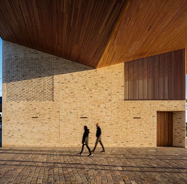Exterior architectural photograph of a large contemporary brick building with a warm, finely slatted timber soffit and matching vertical timber screening. The low, monolithic facade includes a recessed timber entry door, narrow horizontal openings, and a broad brick-paved forecourt. Two pedestrians provide scale.