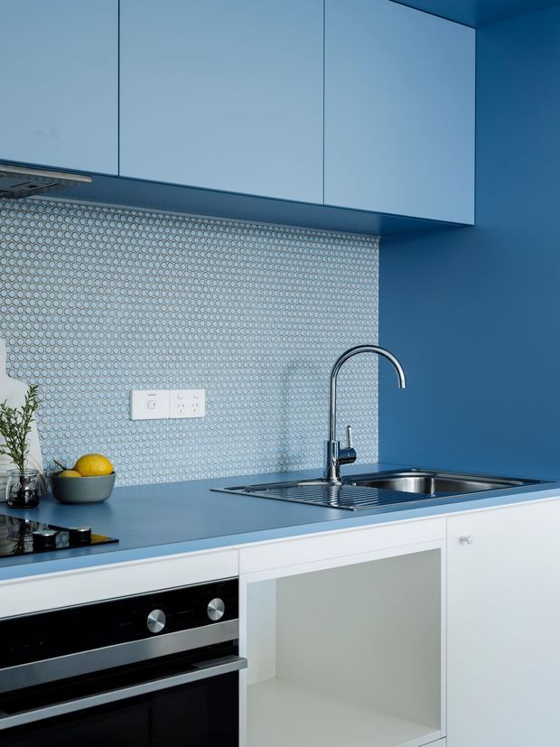 A compact contemporary kitchen in a vivid blue and white palette. The image shows blue upper cabinetry and walls, a blue benchtop, white lower cabinetry, a stainless-steel undermount sink and curved tap, integrated oven, black cooktop, and a small circular mosaic tile splashback. The composition focuses on the kitchen work zone and fixed joinery.