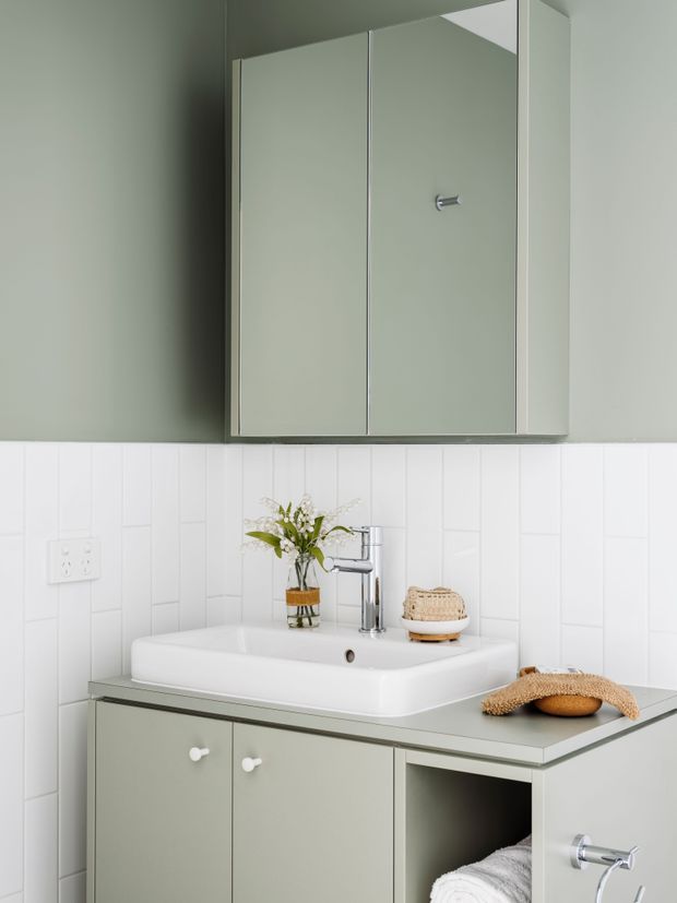 A compact, bright bathroom vanity with a white ceramic basin, chrome mixer tap, pale sage-green built-in cabinetry, and a large mirrored wall cabinet. The walls feature white vertical rectangular tiles with matching grout, while small woven accessories and a simple plant add natural texture.