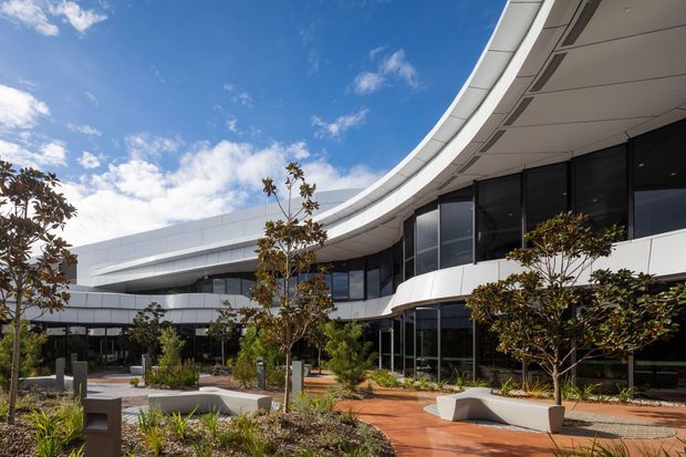 Exterior architectural view of a large contemporary commercial building arranged around a landscaped courtyard. The building has sweeping curved roof and canopy lines, white horizontal cladding, extensive dark-framed glazing, and a paved pedestrian forecourt with integrated angular concrete seating and garden beds. Clear blue sky and mature planted trees frame the scene.