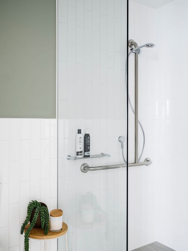 A bright, compact accessible-style shower enclosure with clear glass screens, white vertical rectangular wall tiles, chrome grab rails, a handheld shower on a vertical rail, and a small corner shelf holding toiletries. A muted sage-green painted wall and a small wooden corner stool with a trailing plant and candle add warmth.