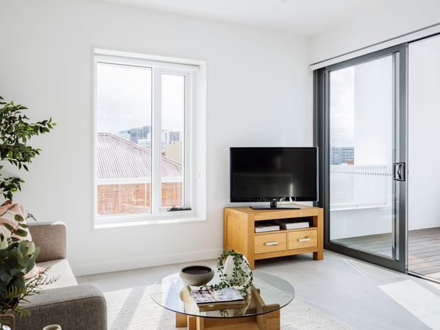 Bright contemporary living room with white walls, large windows, and full-height sliding glass doors opening to a balcony. The room features a grey upholstered sofa, a round glass coffee table, and a mid-tone timber TV console with a flat-screen television. Urban buildings are visible outside.