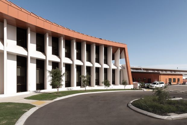 Exterior architectural photograph of a contemporary multi-storey commercial building with a long curved terracotta-orange roof and matching upper facade band. Repetitive white vertical structural bays frame dark recessed windows, creating a strong rhythmic facade. The building fronts a paved driveway and landscaped traffic island, with a lower commercial wing and parked vehicles visible in the background.
