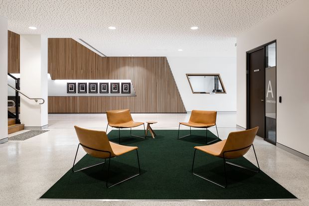 A contemporary commercial reception or waiting area with four tan leather lounge chairs arranged around a small low table on a large dark green rug. The space features polished concrete flooring, white walls, a textured acoustic ceiling, a dramatic angled wall clad in vertical timber slats, framed artwork, a glazed reception opening, and a dark doorway with large lettering. A stairway is partially visible at the left.