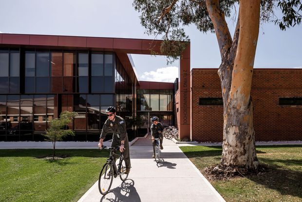 Exterior view of a contemporary institutional building, likely a school, with a glazed dark-framed wing, deep reddish-brown metal fascia, and an adjoining red brick volume. A paved pedestrian path leads through landscaped lawns toward the entrance, with two cyclists in the foreground and a large eucalyptus tree beside the building.