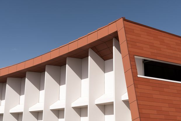 Close-up exterior view of a contemporary flat-roofed building. The facade combines warm terracotta-red horizontal cladding panels with a repeating white projecting structural or sun-shading bay pattern, creating strong geometric rhythm and deep shadow lines. A dark horizontal window opening is visible on the right, beneath a clear blue sky.