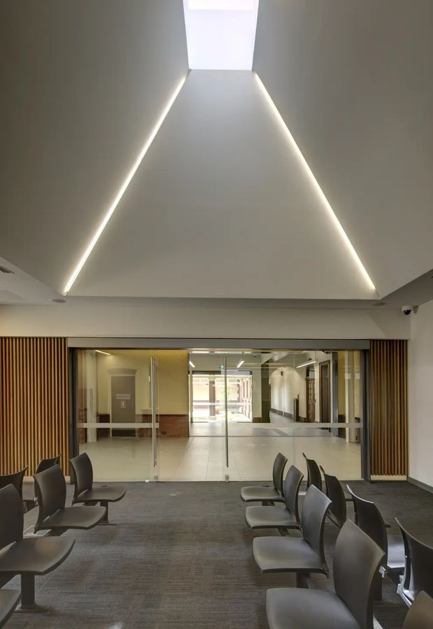 A contemporary commercial interior resembling a reception or waiting area. Rows of dark upholstered chairs sit on grey carpet in the foreground, facing a glazed entry or corridor. Vertical timber slat wall panels frame the opening, while concealed linear LED lighting outlines a large angular recessed ceiling feature and a bright rectangular skylight or ceiling panel. The space beyond the glass has light tiled flooring, reflective surfaces, and additional circulation areas.
