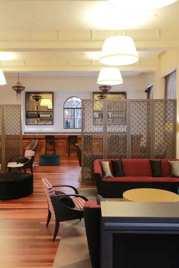 A refined hospitality-style lounge and bar interior with warm timber flooring, a wood-paneled bar, patterned metal room dividers, upholstered red and black seating, and layered pendant lighting. Arched windows and decorative screens add a classic architectural character, while the varied seating zones create a sophisticated, inviting atmosphere.