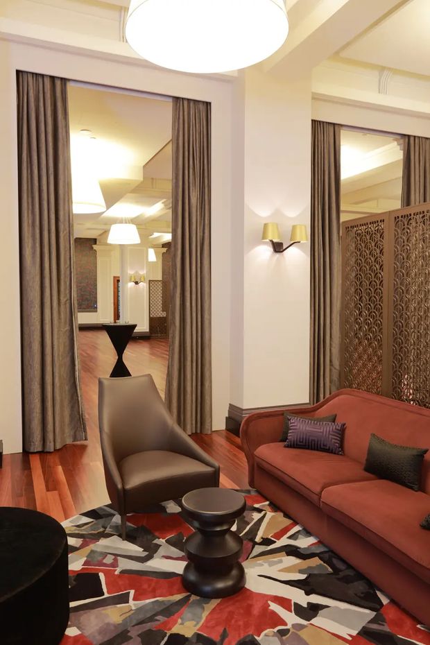 A richly styled living room with a warm, refined interior. The room features polished reddish timber flooring, cream walls with substantial architectural mouldings and columns, tall curtained openings, wall sconces, a large circular pendant light, a rust-red curved sofa, sculptural dark coffee tables, a leather lounge chair, and a bold multicoloured abstract rug. Decorative patterned screens and mirrored openings add depth and an art-deco-influenced character.
