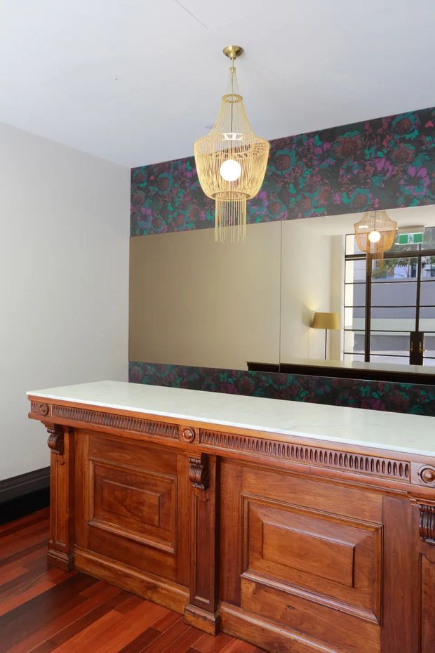 An elegant commercial or hospitality reception area featuring a large ornate dark timber reception counter with a pale stone benchtop, polished timber flooring, and a large wall mirror. A dramatic floral-patterned wall covering frames the mirror, while a sculptural brass-gold pendant light hangs centrally above the counter. The space combines heritage-style joinery with bold decorative detailing and contemporary lighting.