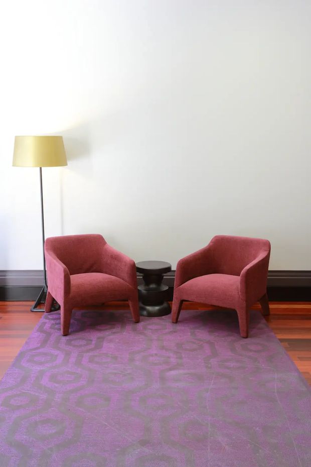 A minimalist seating vignette against a plain white wall, featuring two deep rose upholstered armchairs, a small black round side table, and a slender floor lamp with a yellow shade. The furniture sits on a large purple patterned rug over polished reddish timber flooring, with dark baseboard trim along the wall. The composition is simple and symmetrical with a restrained, gallery-like feel.