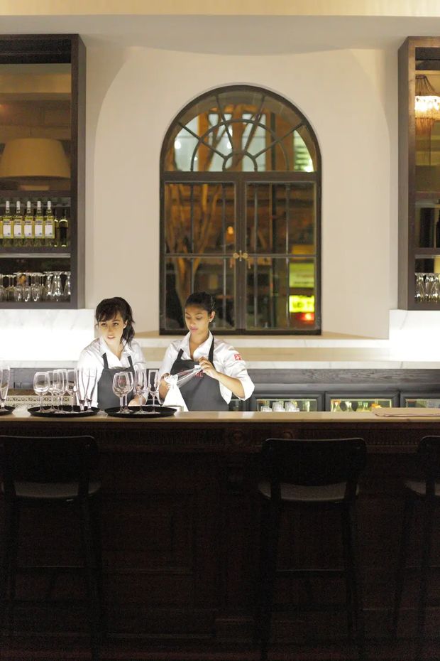 A warmly lit commercial bar interior with a dark timber bar counter and stools in the foreground. Two bartenders stand behind the counter, with glassware and bottles displayed on open shelves. The rear wall features a prominent arched, divided window or glazed opening with decorative metalwork, flanked by built-in display shelving.