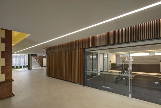 Modern commercial interior lobby or circulation area with a large curved wall clad in vertical timber slats. Full-height glass partitions reveal a contemporary meeting or work area with a long table and chairs. The space has pale tiled flooring, a smooth light ceiling with linear recessed lighting, dark metal window and door frames, and a partially visible brick-and-stone feature wall. A stairway and glazed entrance area extend into the background.
