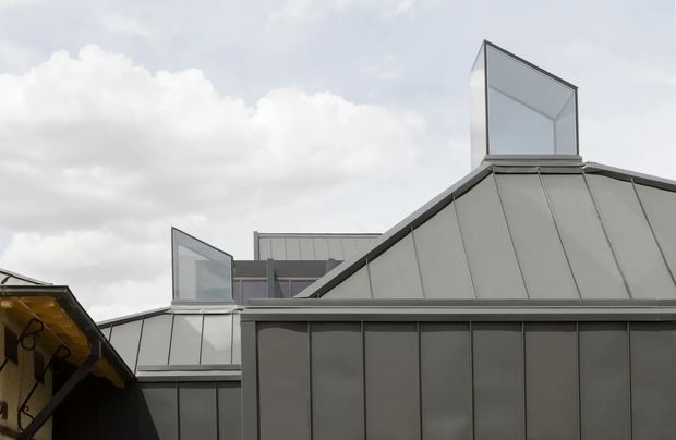 Exterior architectural view of a contemporary building with a sharply pitched, folded roof form clad in grey metal panels. Two raised glazed roof monitors or clerestory structures project above the roofline, with dark-framed vertical glazing and a muted overcast sky. A small portion of an adjacent structure is visible at left.