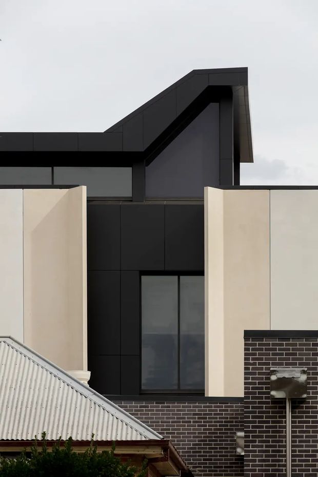 Close-up exterior view of a contemporary multi-storey residential building, featuring a sharply folded dark roof form, black cladding and framing, pale rendered wall planes, tall dark glazing, and contrasting dark brickwork. A corrugated metal roof from an adjacent or lower structure is visible in the foreground.