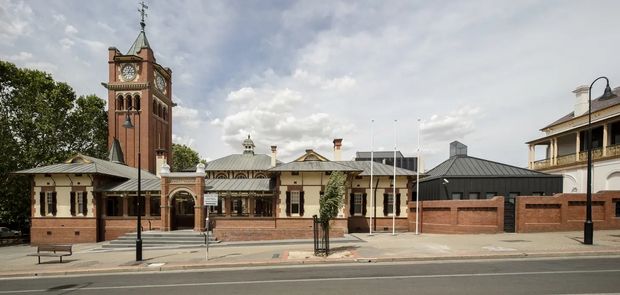 Street-facing view of a historic civic or institutional building complex, dominated by a tall red-brick clock tower with a steep metal spire. The adjoining low-rise buildings feature cream rendered walls, decorative arched detailing, dark metal pitched roofs, brick boundary walls, and traditional verandah elements. A larger multi-storey heritage-style building is visible at right, with a paved street and footpath in the foreground.