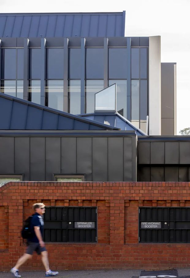Street-level view of a contemporary multi-storey building with dark metal standing-seam cladding, extensive vertical glazing, geometric roof forms and a pale rendered side volume. A lower red-brick boundary wall contains black utility access panels labelled sprinkler booster and fire hydrant booster, with a blurred pedestrian in the foreground.