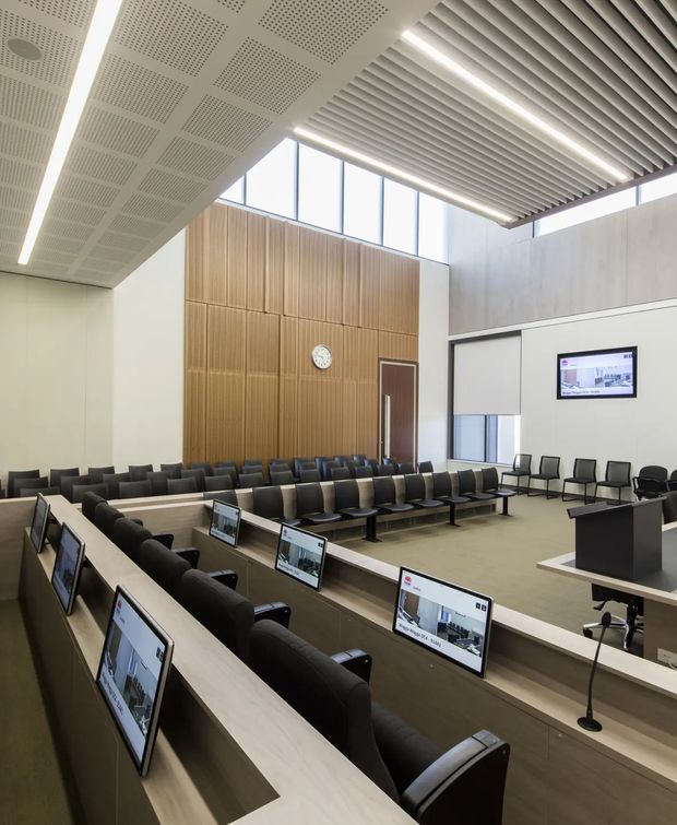 A bright, formal institutional hearing or courtroom-style chamber with tiered rows of built-in desks, black upholstered chairs, individual computer monitors, and a lectern. The room features a tall vertical timber-slat feature wall with a clock and door, pale neutral walls, high clerestory glazing, and a sculptural linear slatted ceiling with integrated strip lighting. The space has a clean, contemporary commercial design.