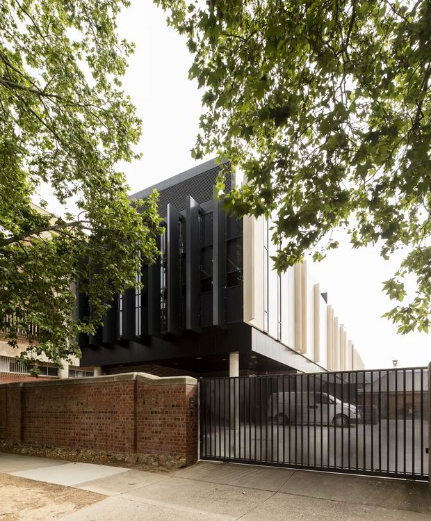 Street-level exterior view of a contemporary multi-storey building framed by mature leafy trees. The building has a strong dark facade with deep vertical fins and screens, contrasting with a lighter side elevation featuring repeated projecting vertical elements. A brick boundary wall, black metal security gate, driveway, and parked vehicle are visible at ground level.
