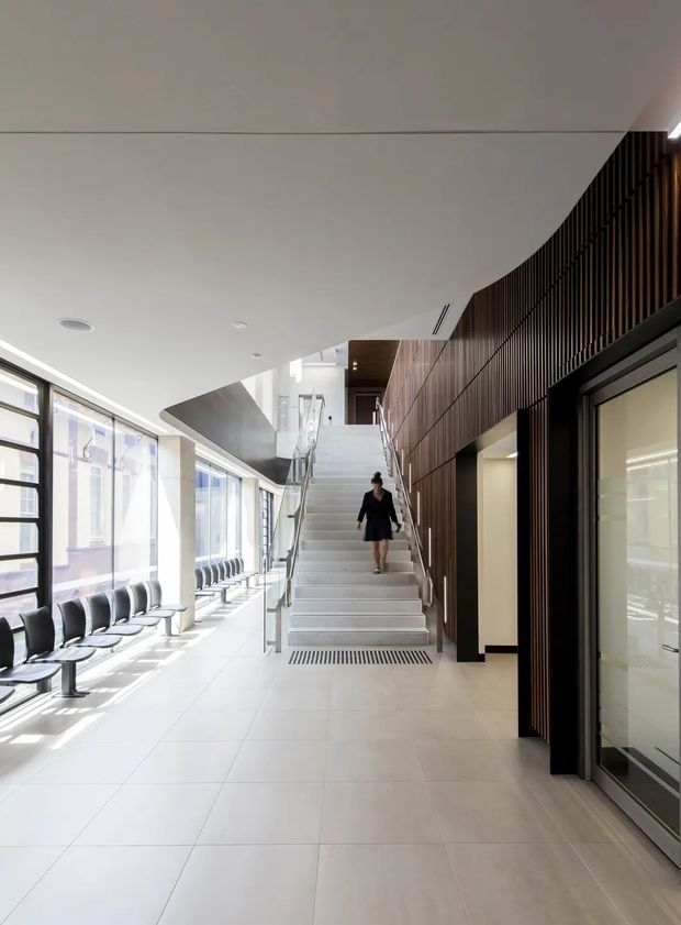 Bright contemporary interior circulation space with a broad tiled floor, a central straight staircase, extensive floor-to-ceiling glazing and rows of waiting chairs along the left side. The right wall features dark timber vertical slatted panelling and dark-framed glazed doors, while a curved white ceiling and glass stair balustrade create a clean, modern institutional or commercial feel.