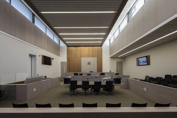 A large contemporary council or courtroom-style chamber with tiered seating, a central meeting table, integrated desks, wall-mounted screens, pale grey architectural finishes, and a warm timber feature wall. The room has a high linear slatted ceiling with recessed lighting and clerestory glazing along both sides, creating an institutional yet refined interior.