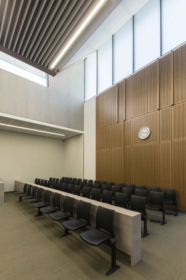 A contemporary institutional lecture hall or auditorium with tiered rows of black seats and long pale desks. The room features a tall double-height volume, large frosted clerestory windows, a dramatic slatted timber ceiling with integrated linear lighting, and a full-height ribbed timber feature wall with an analog clock. The palette is restrained in warm grey, pale timber, and black, creating a refined, modern academic or commercial interior.