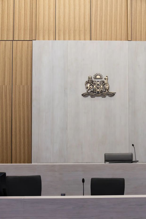 A formal courtroom or tribunal chamber with pale grey stone or concrete wall panels, warm vertical timber panelling, a raised bench with microphones, black leather seating, and a prominent heraldic crest mounted on the rear wall. The space has a restrained institutional and contemporary appearance.