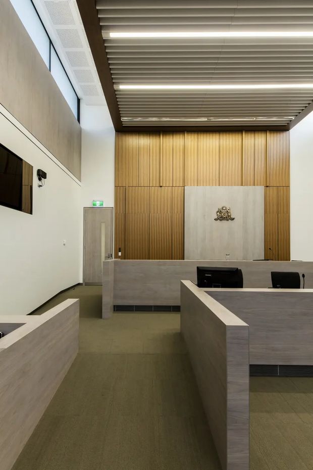 A modern commercial reception or public-service lobby with a central reception desk, pale grey wood-look counters, vertical timber wall panelling, a large light stone or concrete feature panel bearing a crest, and a suspended linear slatted ceiling with integrated strip lighting. The space is tall, clean-lined, and institutional in character, with a wall-mounted display screen and an exit door.