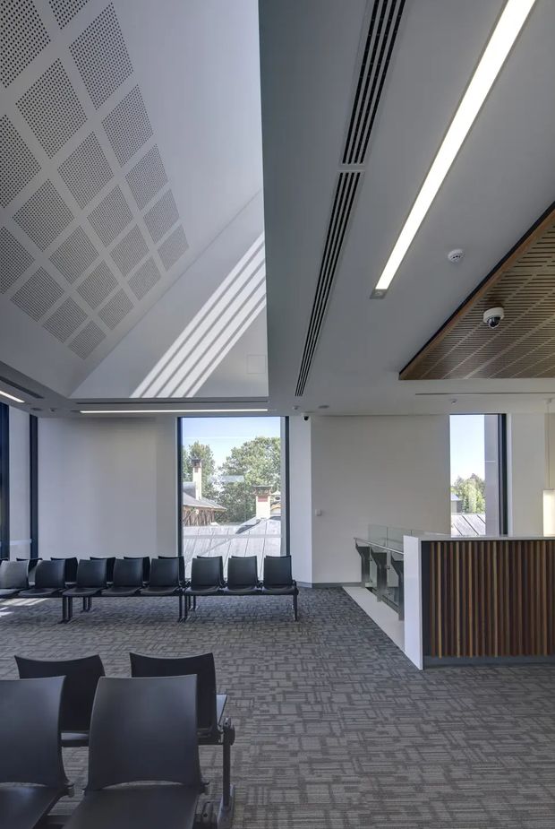 A bright contemporary commercial waiting or reception area with rows of dark upholstered chairs, grey patterned carpet, large windows, and a reception counter with vertical timber slat detailing. The space features a high white ceiling with perforated acoustic panels, linear air-conditioning grilles, recessed strip lighting, and a glazed stair or balustrade area. The setting appears institutional or office-like, with clean modern detailing and abundant daylight.